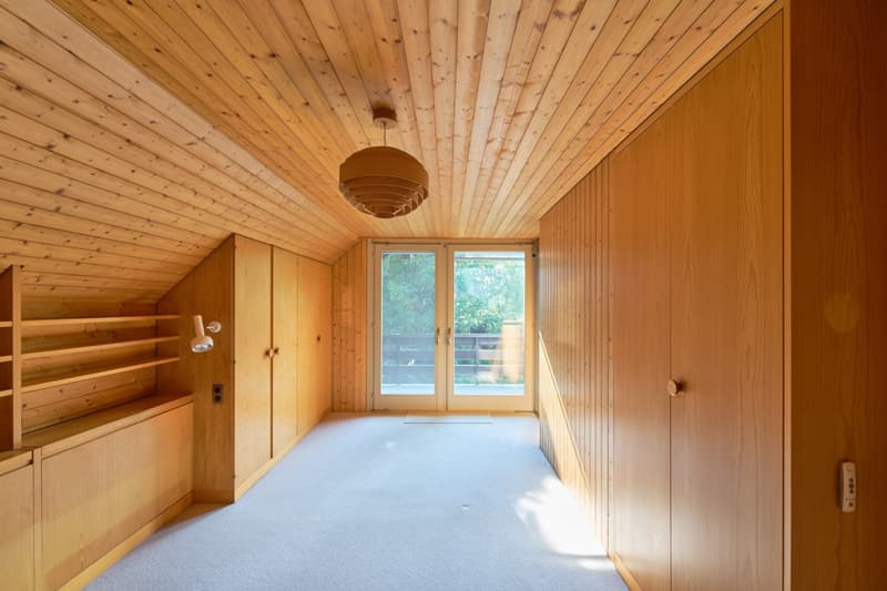 Spacious room with wooden paneled walls and ceiling, large windows providing natural light, built-in shelving, and a pendant light fixture.
