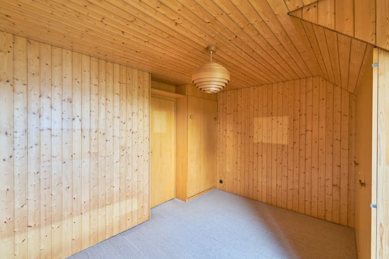This image shows a room with wooden paneled walls and ceiling, a light fixture in the center, and a bare floor. The room appears to be empty and has a simple, minimalist design.
