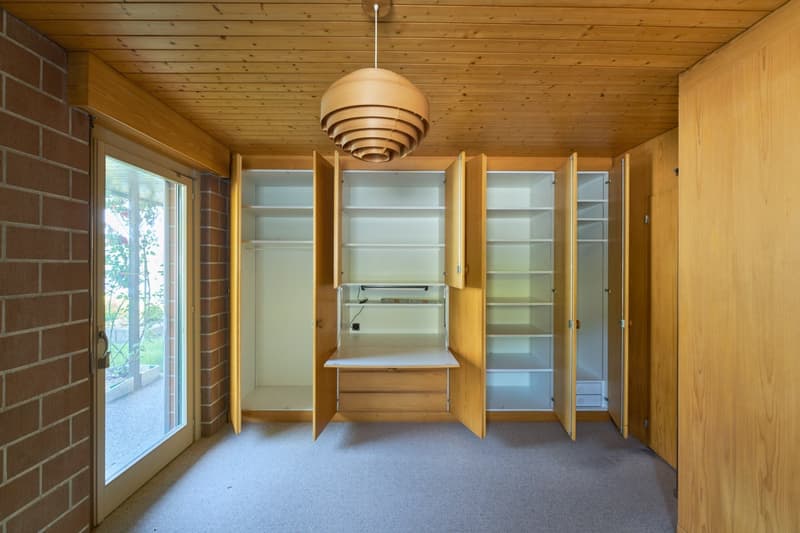 This image shows a room with a wooden ceiling and walls, as well as a large built-in storage unit with shelves and cabinets. There is a window that provides natural light, and a pendant light fixture hanging from the ceiling. The room appears to be a bedr
