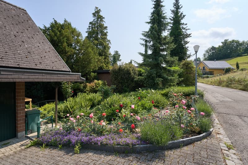 2-story house with a tiled roof, surrounded by lush greenery and a well-maintained garden. The property features a paved walkway, a lamp post, and a variety of colorful flowers and plants.