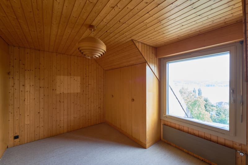 This image shows a room with wooden paneled walls and ceiling, a large window providing a view of the outdoors, and a light fixture hanging from the ceiling.