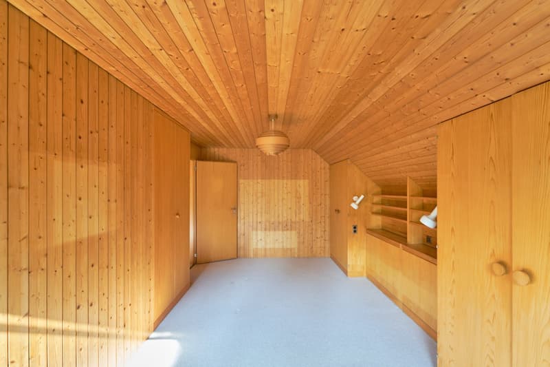 The image shows the interior of a wooden sauna room. The walls and ceiling are made of natural wood panels, creating a warm and cozy atmosphere. The room has a wooden bench along the walls, and there is a light fixture hanging from the ceiling. The floor