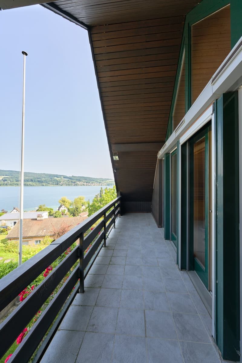 Spacious balcony with wooden ceiling, large windows, and a scenic view of the lake and surrounding landscape.