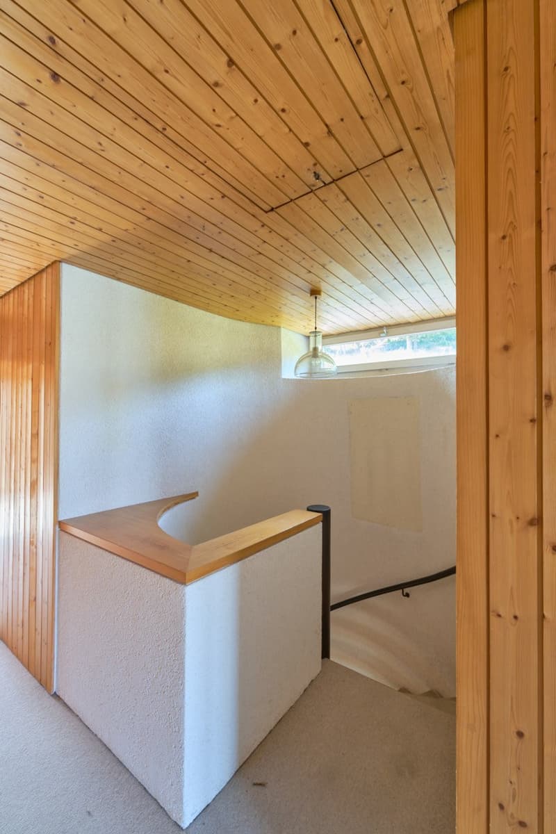 The image shows a small room with a wooden ceiling and walls. There is a counter-like structure with a wooden top, and a light fixture hanging from the ceiling. The room appears to be a simple, minimalist space with a clean and natural aesthetic.