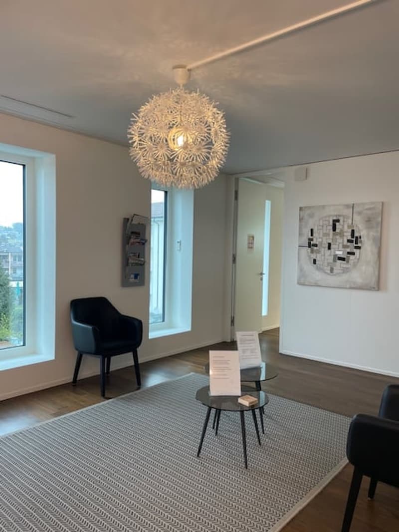 Living room with white walls, wooden floor, large windows, chandelier, two black chairs, two tables, rug, and a painting on the wall