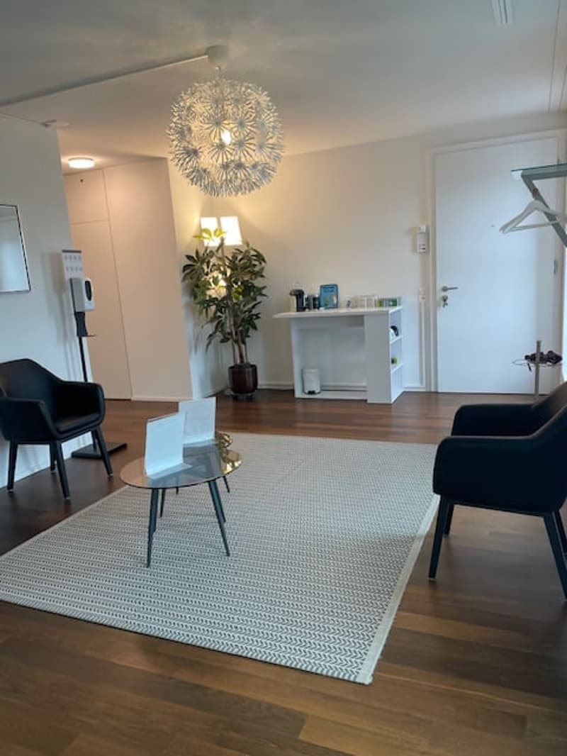 Living room with wooden floor, white walls, chandelier, coffee table, chairs, rug, plant, white desk, and a white door.