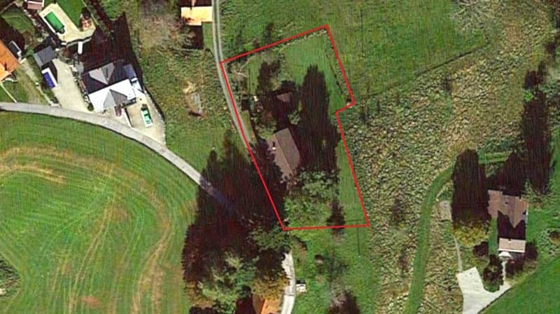 Large open grassy area, bordered by trees, house, buildings on the left side, dirt driveway