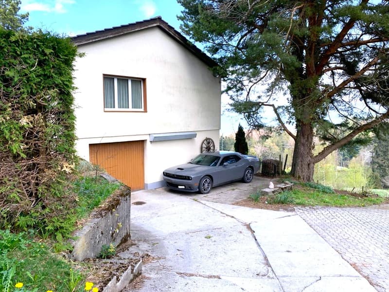 2-story white house with wooden garage door, surrounded by trees and greenery, with a paved driveway leading to a parked car