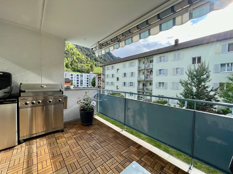 Balcony with wooden floor, stainless steel grill, potted plant, metal railing, awning, and a view of the mountains and buildings.