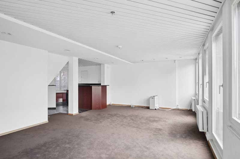 Large open-plan living space with white walls, grey carpeted floor, and a wooden cabinet or kitchen unit visible in the background. The room appears to be in the process of renovation or furnishing, with some equipment or materials visible on the floor.