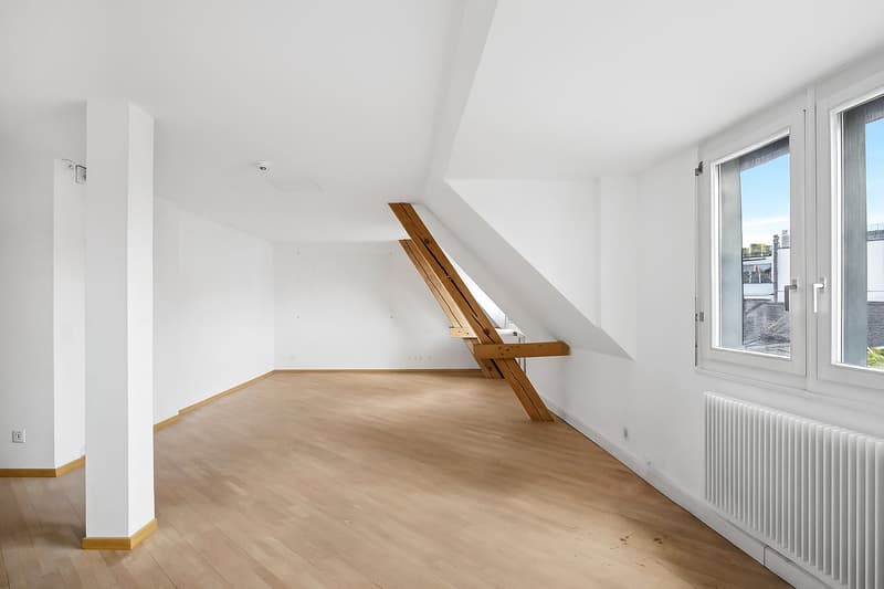 Spacious attic room with white walls, wooden floors, and a large window providing natural light. The room features a wooden staircase or ladder structure against the wall.