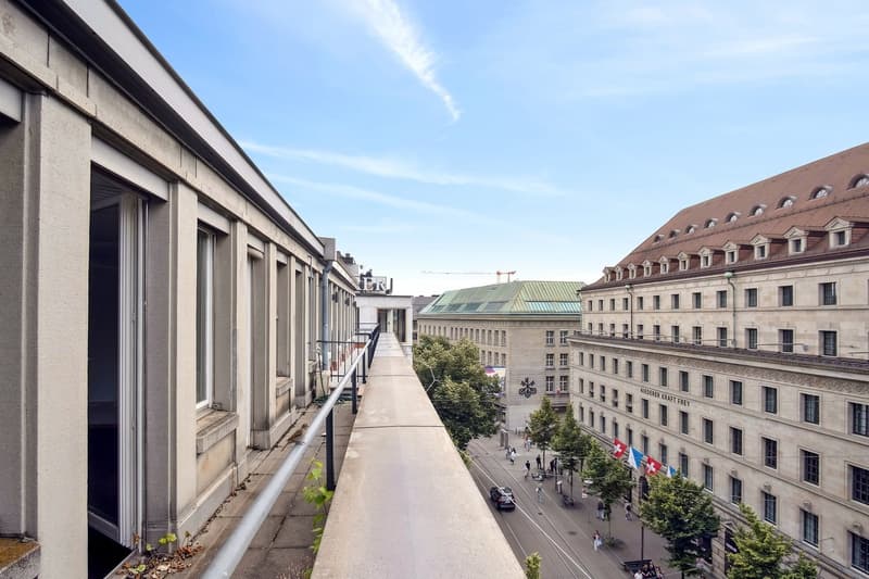 The image shows a view of a city street with a mix of modern and historic buildings. There is a raised walkway or bridge running along the side of the buildings, providing a vantage point to overlook the street below. The buildings have a mix of architect