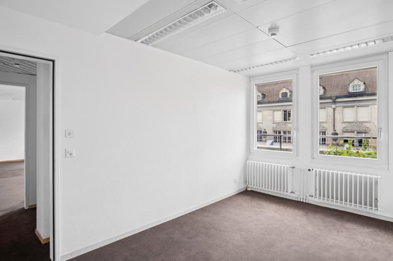 Spacious office room with large windows overlooking a building exterior, white walls, gray carpeted floor, and a radiator heating system.