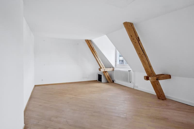 Spacious attic room with slanted ceilings, wooden beams, and hardwood flooring. The room has a large window providing natural light. There is a radiator visible, indicating the room is heated.