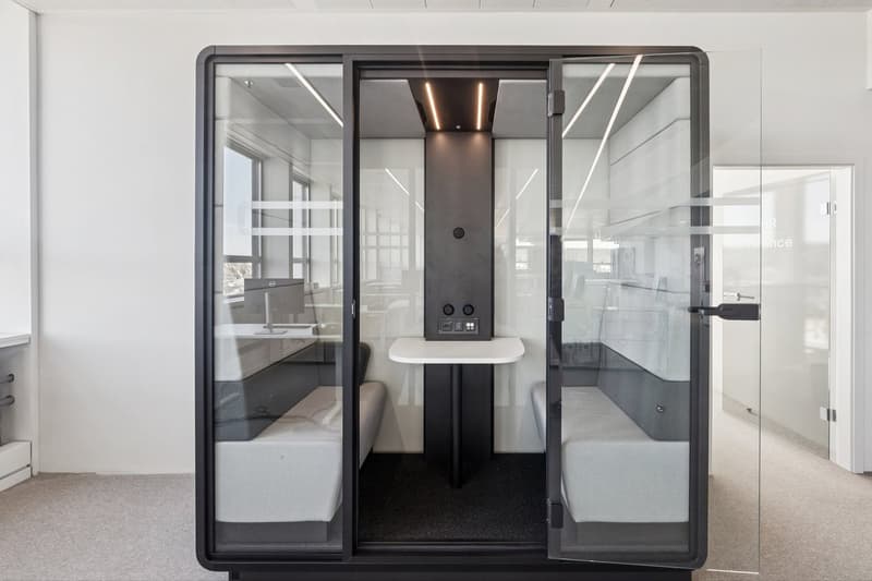 A small meeting room with glass walls, two white benches, a central table, and a black central column with controls.