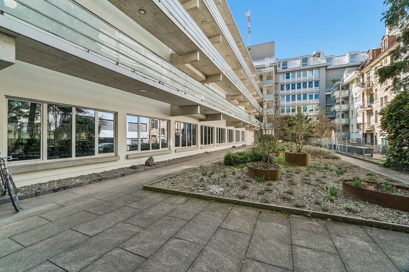 Modern building with glass windows, concrete balcony, paved walkway, small garden with plants