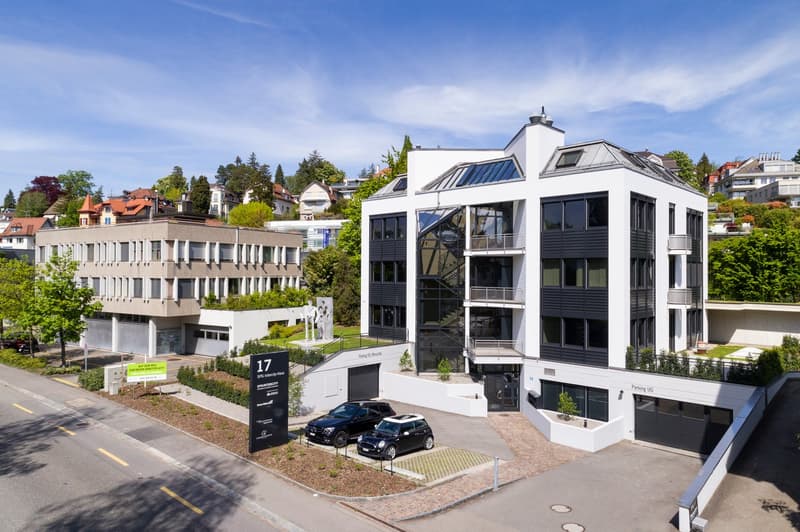 Modern white building with glass windows, two cars parked in front, green plants, and trees in the area