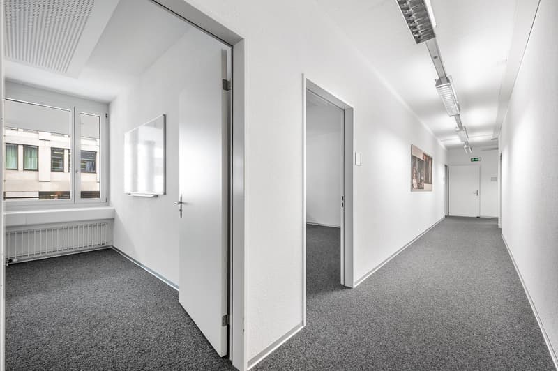 Empty room, white walls, gray carpet, large windows, radiator, two doors, hallway