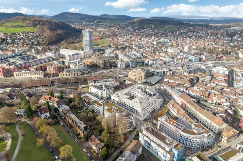 Aerial view of a city with many buildings, roads, and greenery. Mountains in the distance.