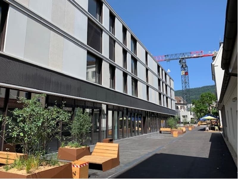 A multi-story commercial or office building with a modern, glass and steel facade. The building has multiple floors and windows, and there is a construction crane visible in the background, indicating ongoing construction or renovation work. The ground le
