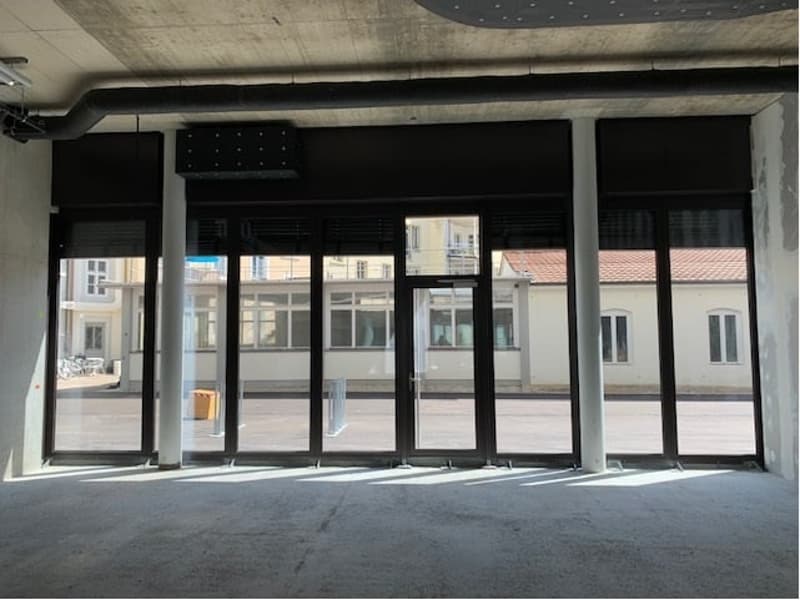 The image shows a large commercial space with floor-to-ceiling windows overlooking a city street. The space appears to be unfinished, with exposed concrete walls and ceiling. The windows provide a clear view of the surrounding buildings.