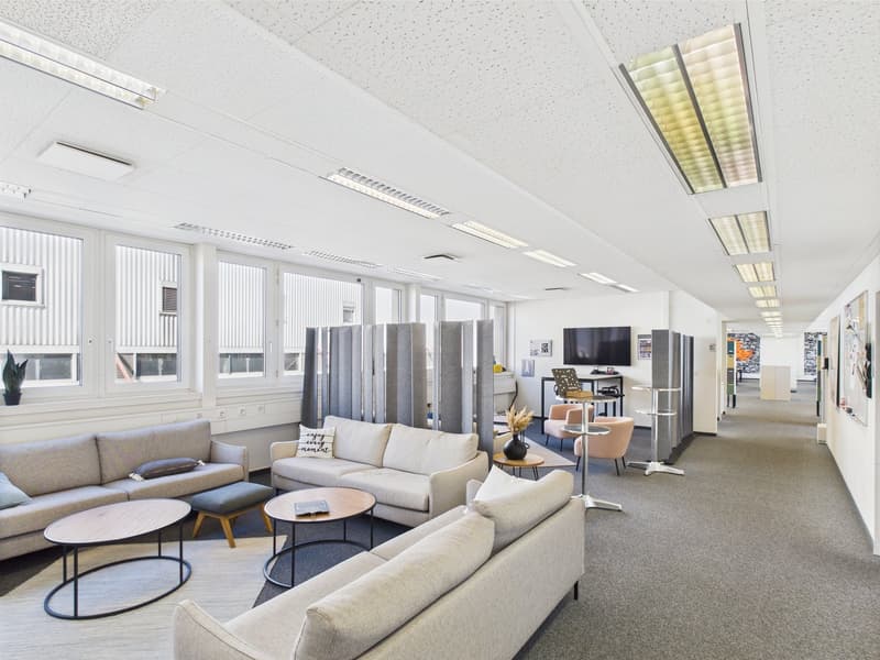 The image shows a modern and open office space with a lounge area. The space features comfortable sofas and armchairs, coffee tables, and a TV mounted on the wall. The ceiling has recessed lighting fixtures and the walls are lined with windows, creating a
