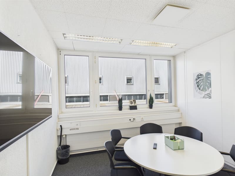 The image shows a modern office space with large windows, white walls, and a round table with chairs. There is a desk with a computer and other office equipment visible. The room has a clean and professional appearance.