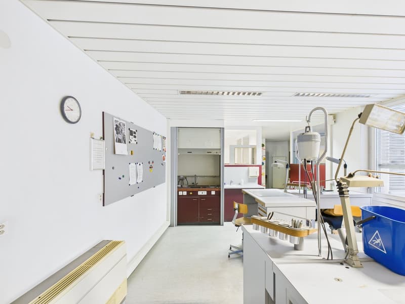 Laboratory space with white walls, white ceiling, clock, noticeboard, worktables, equipment, chairs, trash bin