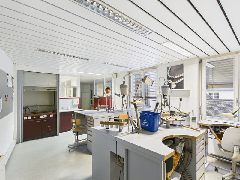 white tiled walls, wooden chairs, desks with tools, sink, cabinets, windows with blinds, bucket, framed picture