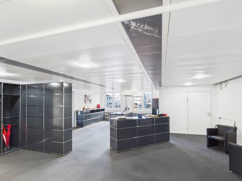 Modern office space with white walls, high ceiling, black cabinets, gray carpet, large windows, open space, chairs, reception desk, desk, monitor, black shelves, picture frame, overhead lights.