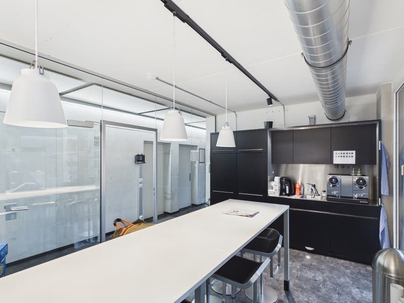 Open plan room, white ceiling, grey floors, black cabinets, white countertops, hanging lamps, large table, stools, coffee machine, microwave