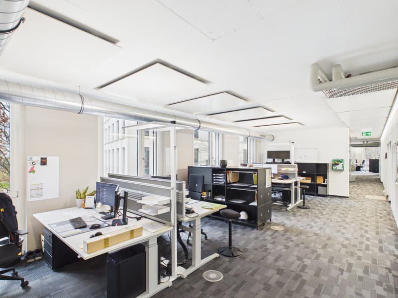 Modern office setup with white desks, adjustable height tables, various office equipment and chairs, large windows