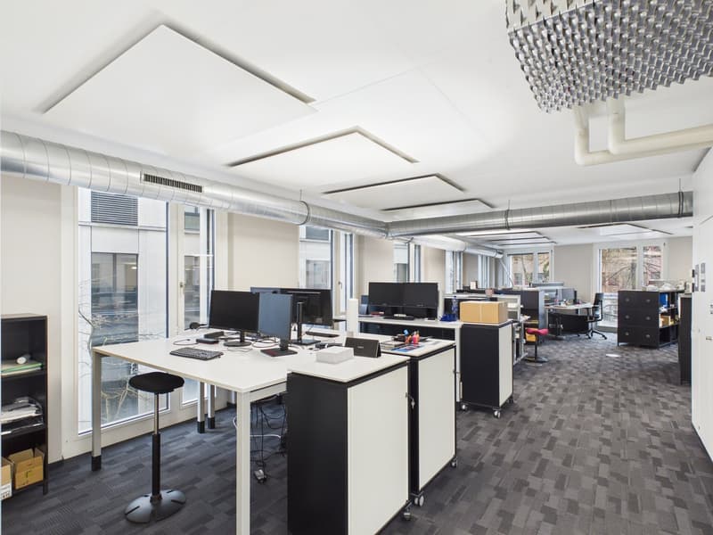 Open space office with white desks, black stools, large windows, and computer monitors.