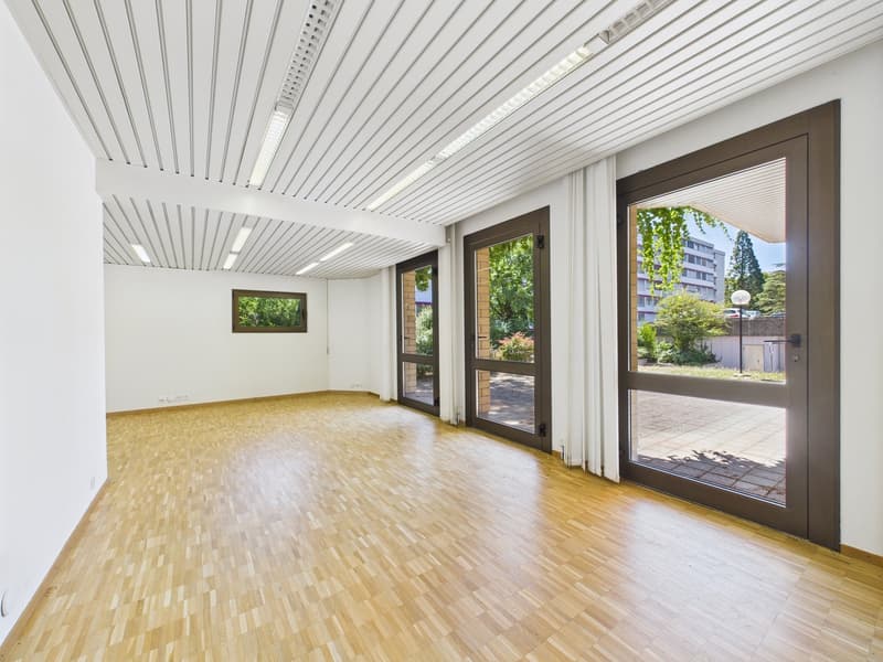 Large open space with high ceilings, wooden flooring, and large windows overlooking a garden area. The room has a modern and minimalist design with a clean, bright atmosphere.