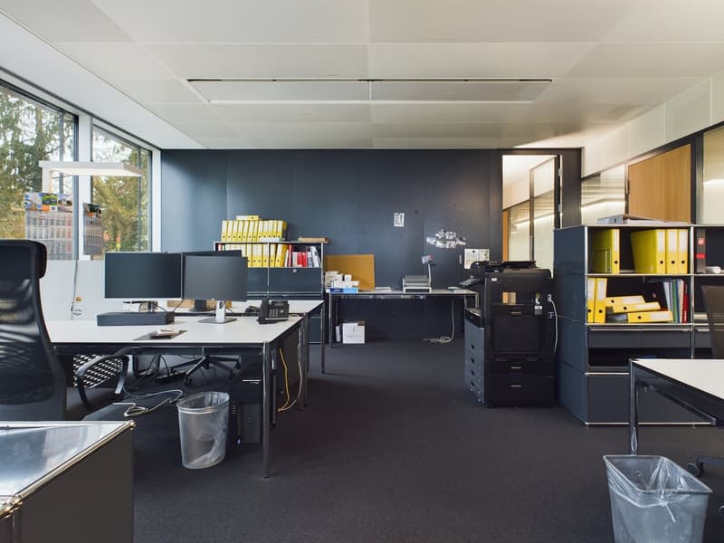 Modern office space with black and white furniture, multiple computer monitors, shelves with binders and folders, and a printer. The room has large windows providing natural light and a dark blue accent wall.