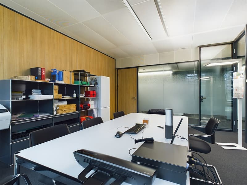 The image shows an office space with a large white desk, a computer, and various office supplies and equipment. The walls are lined with wooden panels, and there is a storage unit with shelves containing various items. The room also has large glass window