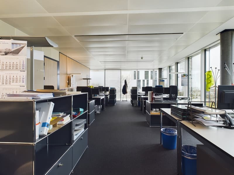 Open office space with cubicles, desks, and storage units. The ceiling has exposed beams and the walls are mostly glass, allowing natural light to enter the space. There are various office equipment and supplies visible, including a dishwasher.