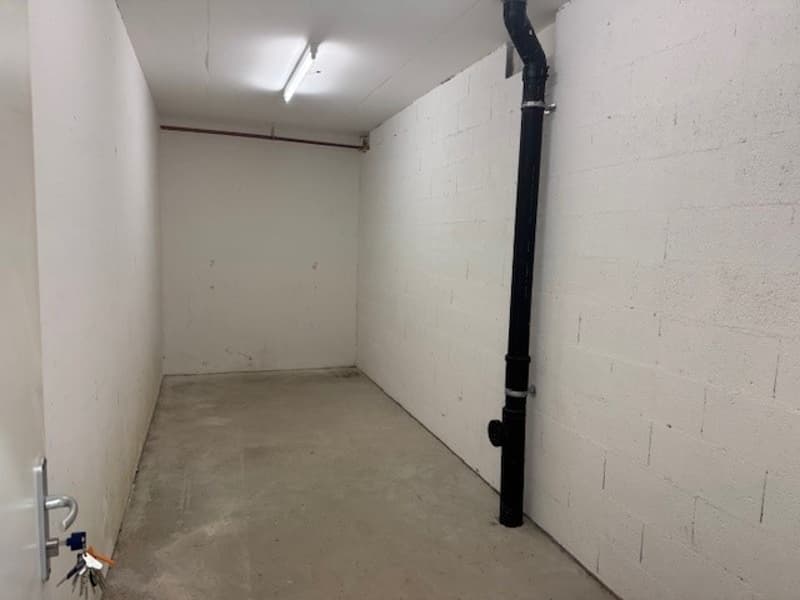 The image shows a small, empty storage room with concrete floors and white walls. There is a large pipe or conduit running along one of the walls.