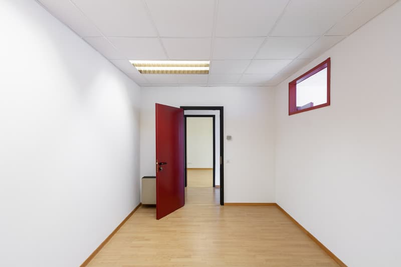 Empty room with wooden floor, white walls, red door, fluorescent ceiling light, a window, and an electrical outlet.