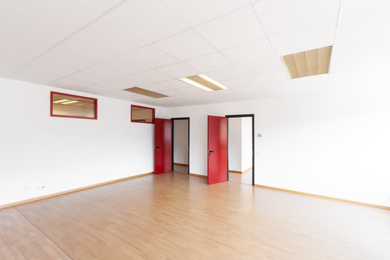 Empty room with white walls, wooden floor, red doors, and small windows.