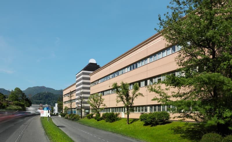 two-story building, rectangular shape, white and beige exterior, multiple windows, surrounded by trees and bushes, road in front