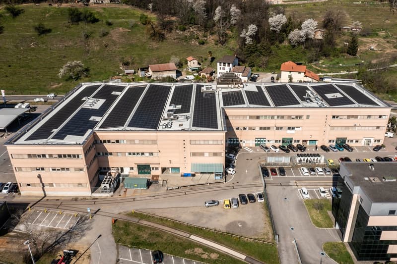 Two-story building, brown exterior, multiple windows, solar panels on roof, parking lot with many cars