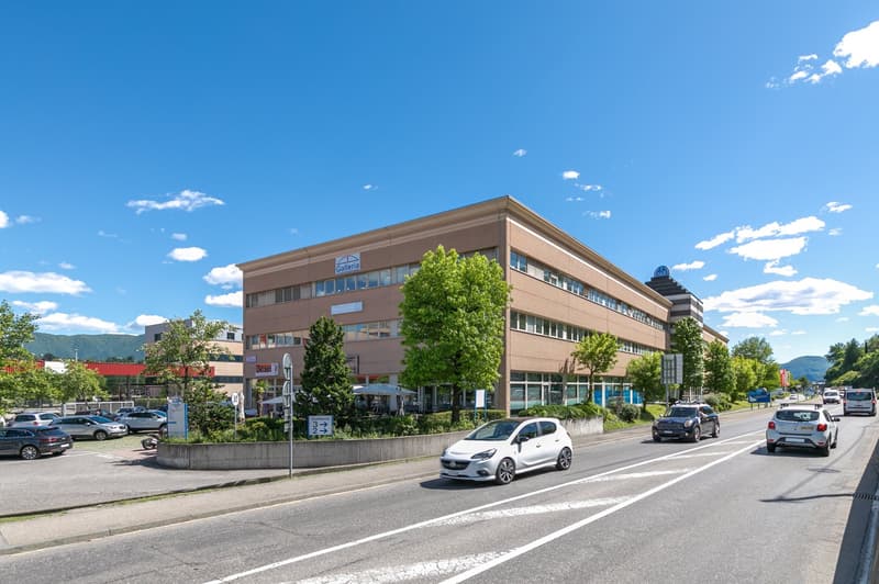 An office building with a car park and greenery outside, situated beside a road with cars driving by.