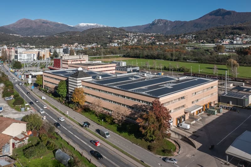 Large multistory building with solar panels, parking lot in front, train tracks, mountains in background.