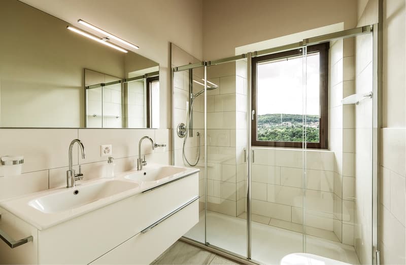 white bathroom with double sink, glass walk-in shower, mirror, electrical outlet, and soap dispenser