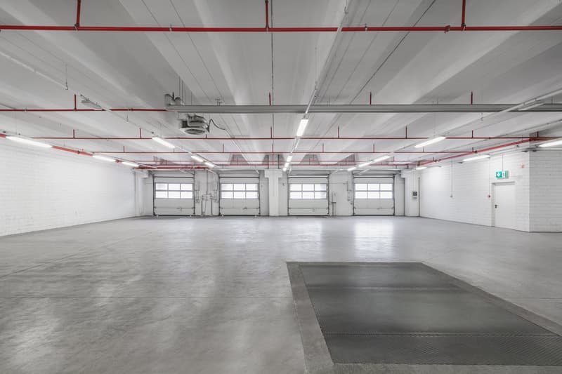 Empty parking lot with white walls, ceiling with pipes, 4 garages, emergency door on right, square gray mat
