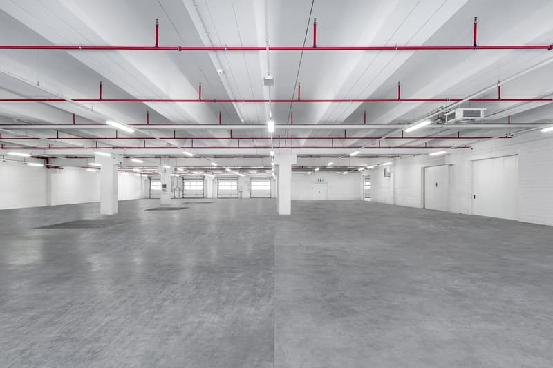 large open space, exposed beams, white and grey walls, multiple garage doors, grey concrete floor, fluorescent ceiling lights