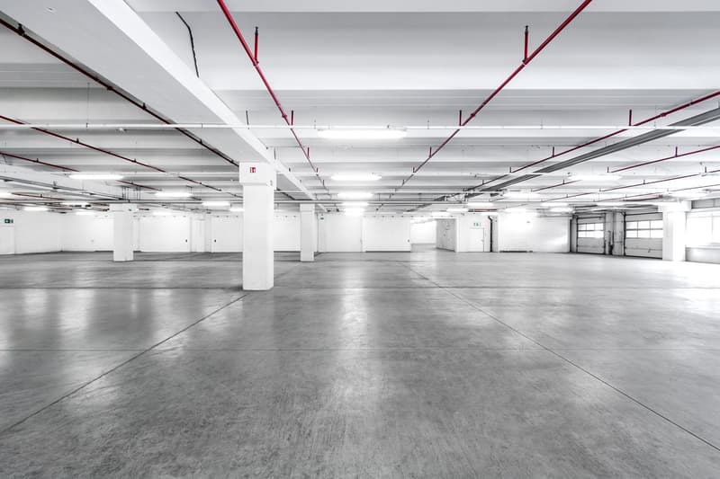 Large empty warehouse with white ceiling, concrete floor, metal beams, columns, roll up doors.