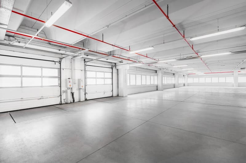 Empty large commercial garage with overhead lighting, roll-up doors, concrete floor, and red piping.