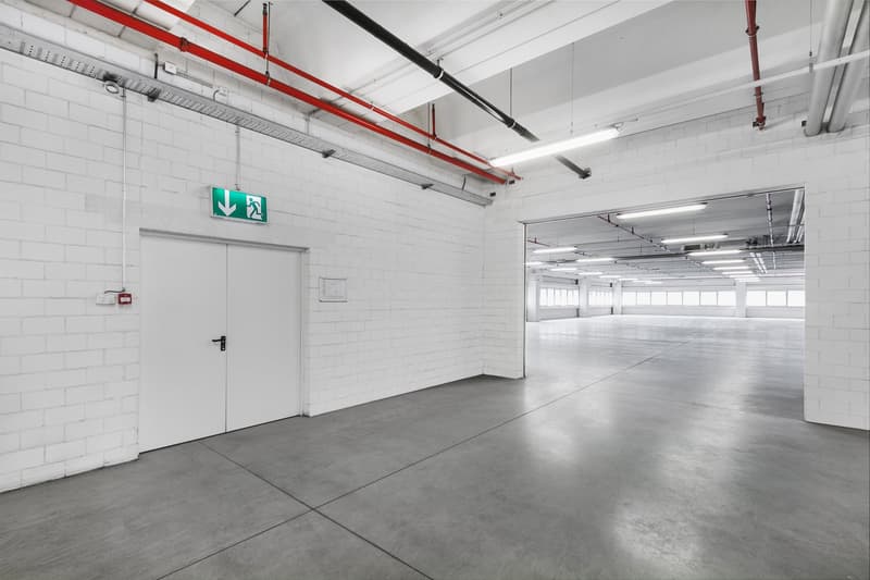 White brick walls, gray concrete floor, emergency exit sign, ceiling with red pipes and lights